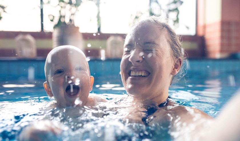 Mother and her nine month old baby boy at his first swimming lesson making selfie in the swimming pool. Smiling, enjoying water, making bubbles and looking at camera. Mother is holding him and helping him to swim.