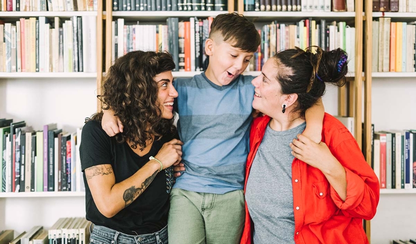 Playful boy standing with arm around women. Happy mothers are looking at affectionate son. They are spending leisure time at home.