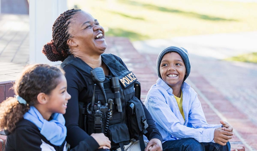 Community policing - a female police officer is conversing with a boy and his sister, sitting side by side on steps outside a building, laughing. The policewoman is African-American, in her 40s. The children are 8 and 7 years old, mixed race African-American and Caucasian.