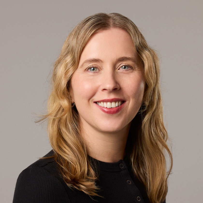 Frances has blonde, shoulder-length wavy hair. Frances is wearing a black button-up jumper.