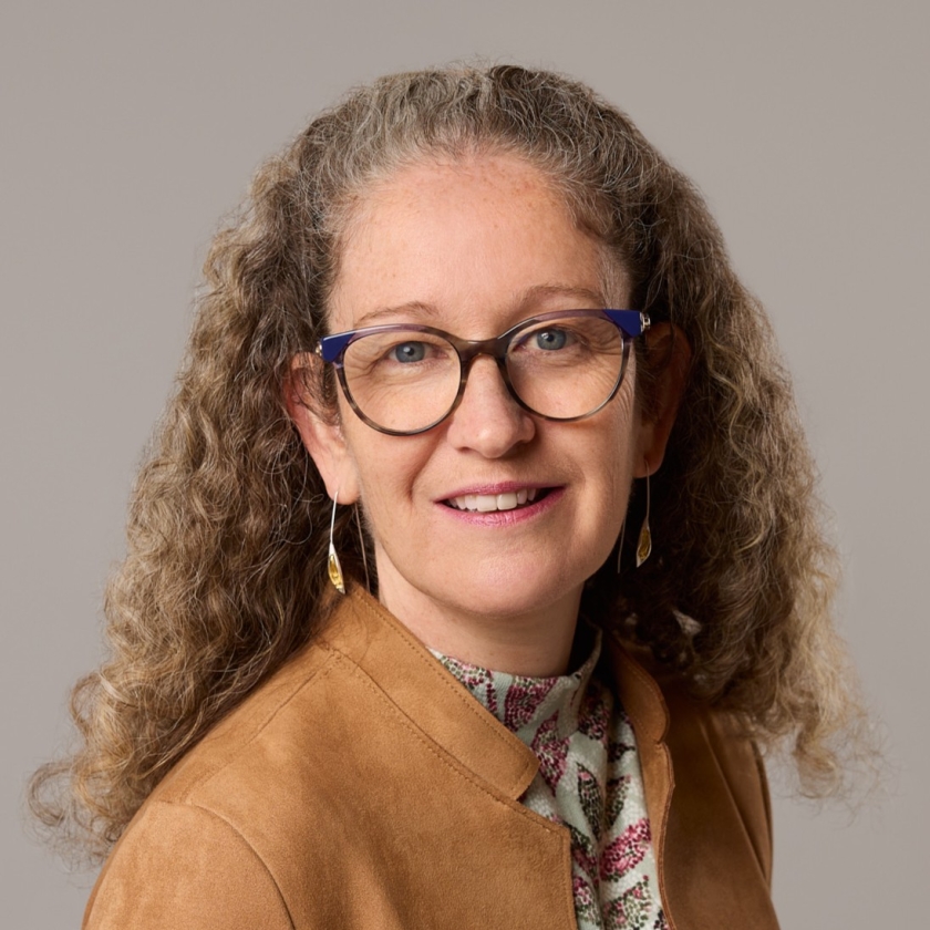 Karen has honey coloured, curly hair. Karen wears blue spectacles, a tan leather jacket and floral shirt.