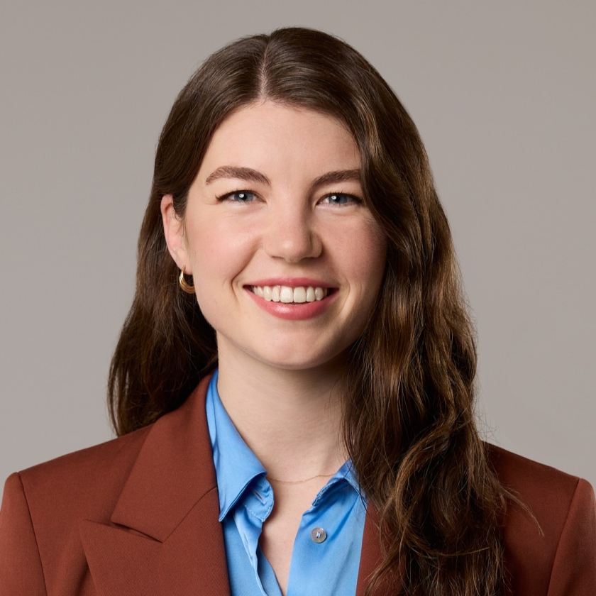 Kate has long, wavy brown hair. Kate is wearing a light blue shirt underneath a terracotta coloured blazer.