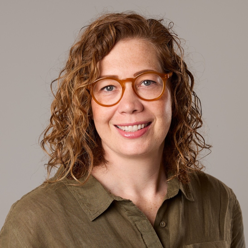 Tamara has shoulder-length, curly light-brown hair. Tamara is wearing dark orange spectacles and a khaki coloured button up shirt. 