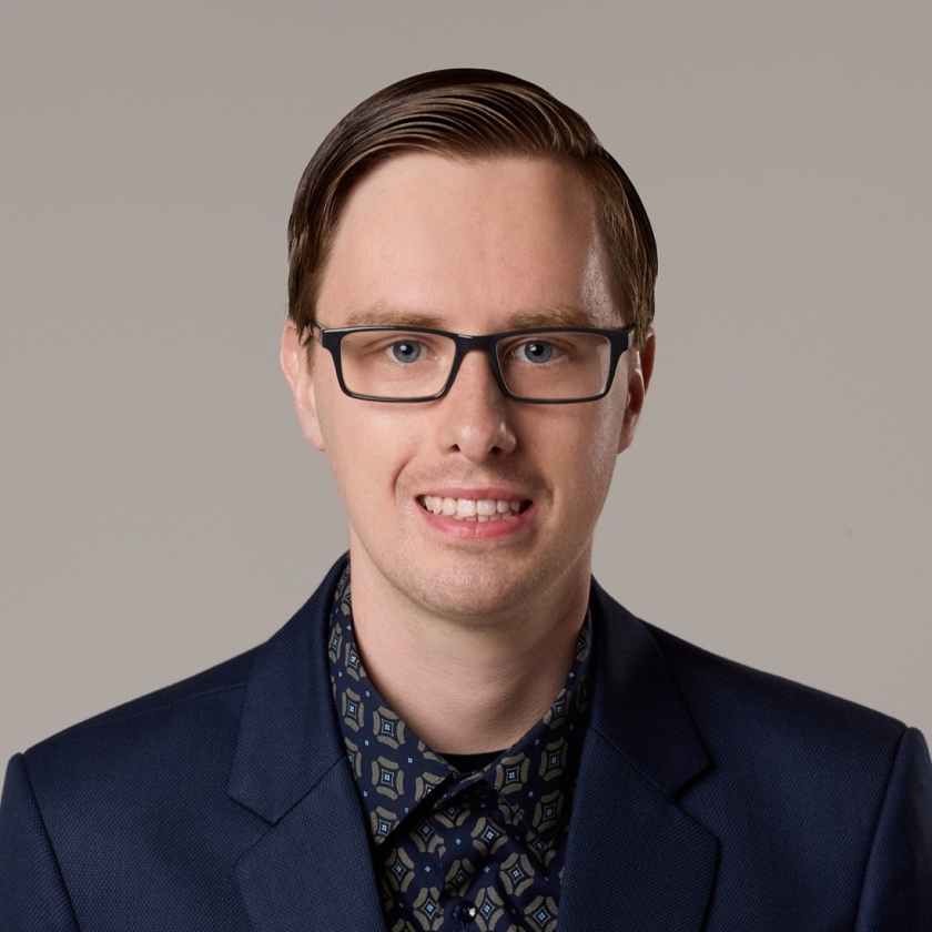 Julian has short, dark brown hair. Julian is wearing black spectacles, a navy blue suit jacket and blue checker button-up shirt.