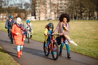 Three generation family walking through a public park. The little boy is cycling through the park with his mothers assistance while she talks to her mother in-law.